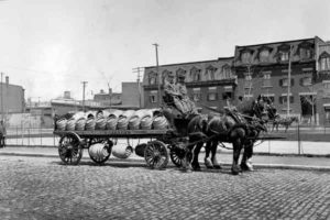 La photo en noir et blanc montre un chariot à quatre roues rempli de tonneaux et tirées par deux chevaux avec deux livreurs.