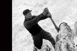 The black and white photo shows a winter scene where a man grips a block of wood with a log hook.