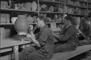 Photographie présentant des hommes travaillant dans un atelier de céramique. Derrière eux se trouvent des pièces non finies.
